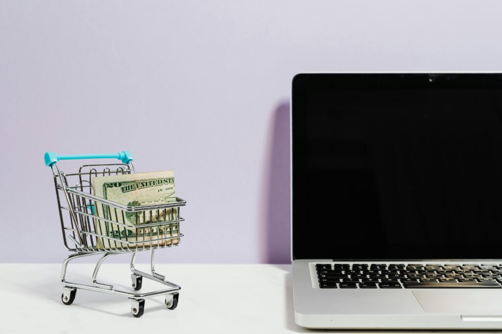 analiza SWOT dla sklepu – wózek zakupowy z pieniędzmi obok laptopa symbolizujący analizę mocnych i słabych stron oraz szans i zagrożeń w biznesie e-commerce